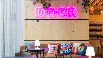 Un espace intérieur accueillant, avec des sièges confortables et des tables, agrémenté d'un néon lumineux sur un mur en bois, sur lequel on peut lire le mot « DOCK ».