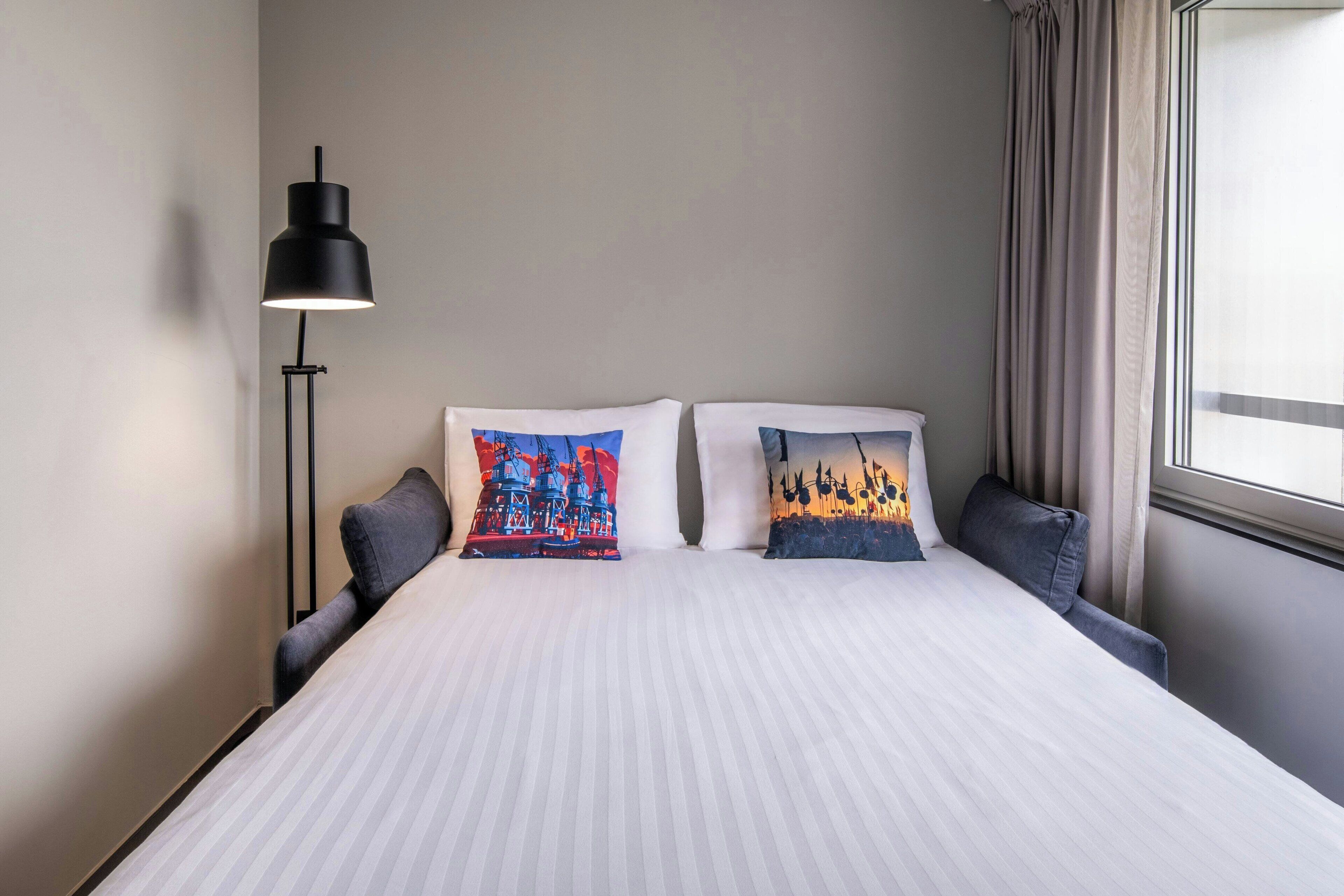 A neatly made bed with striped bedding, two pillows with colorful designs, a black floor lamp, and a window with curtains.