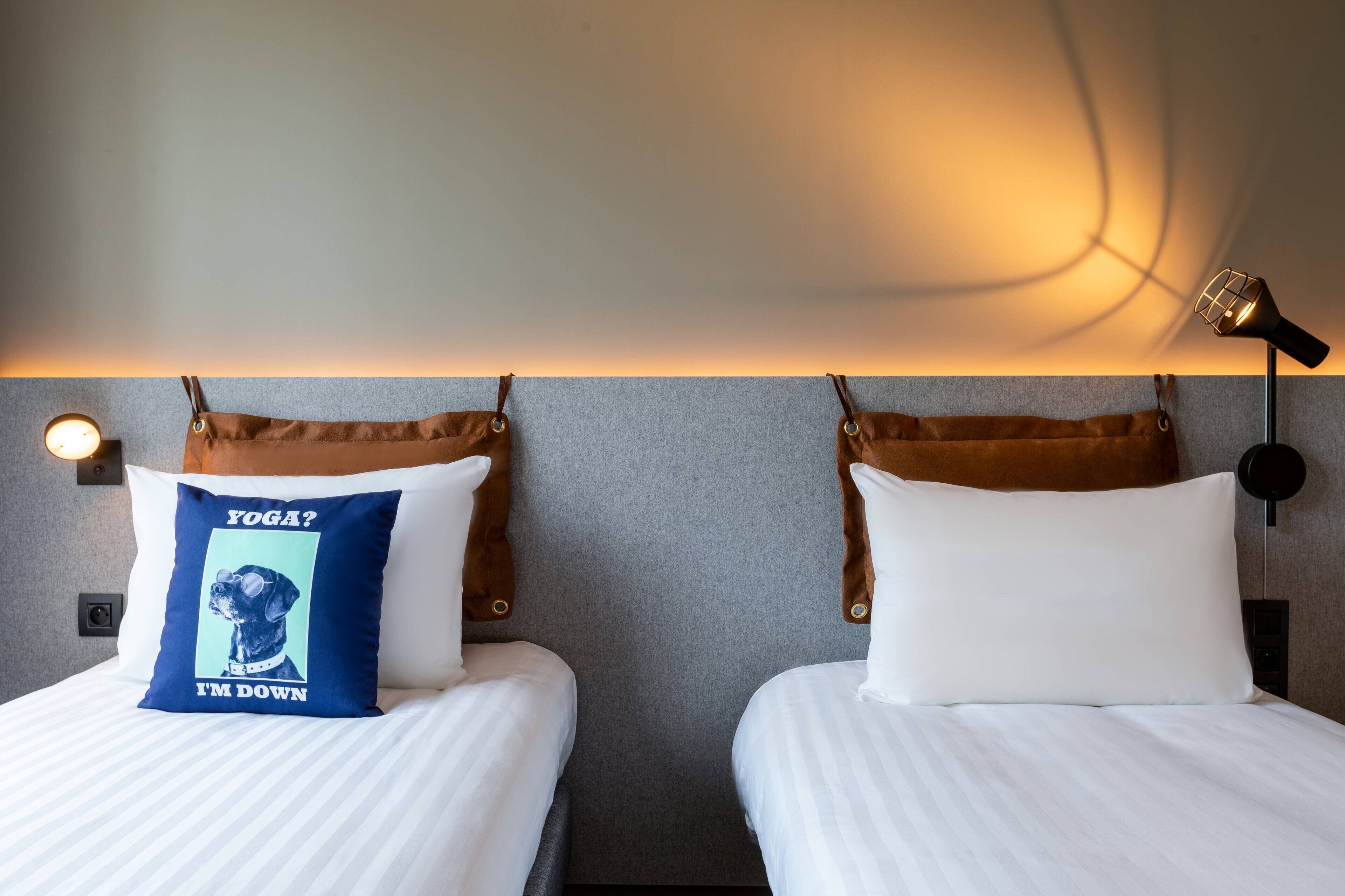 Quarto de hotel com duas camas, cada uma com travesseiro e cabeceira. Há uma luminária na parede e uma tomada.