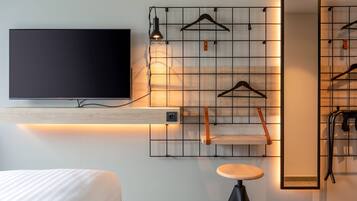 A modern hotel room with a flat-screen TV mounted on a wooden shelf, a wall-mounted mirror, and a seating area with a stool.