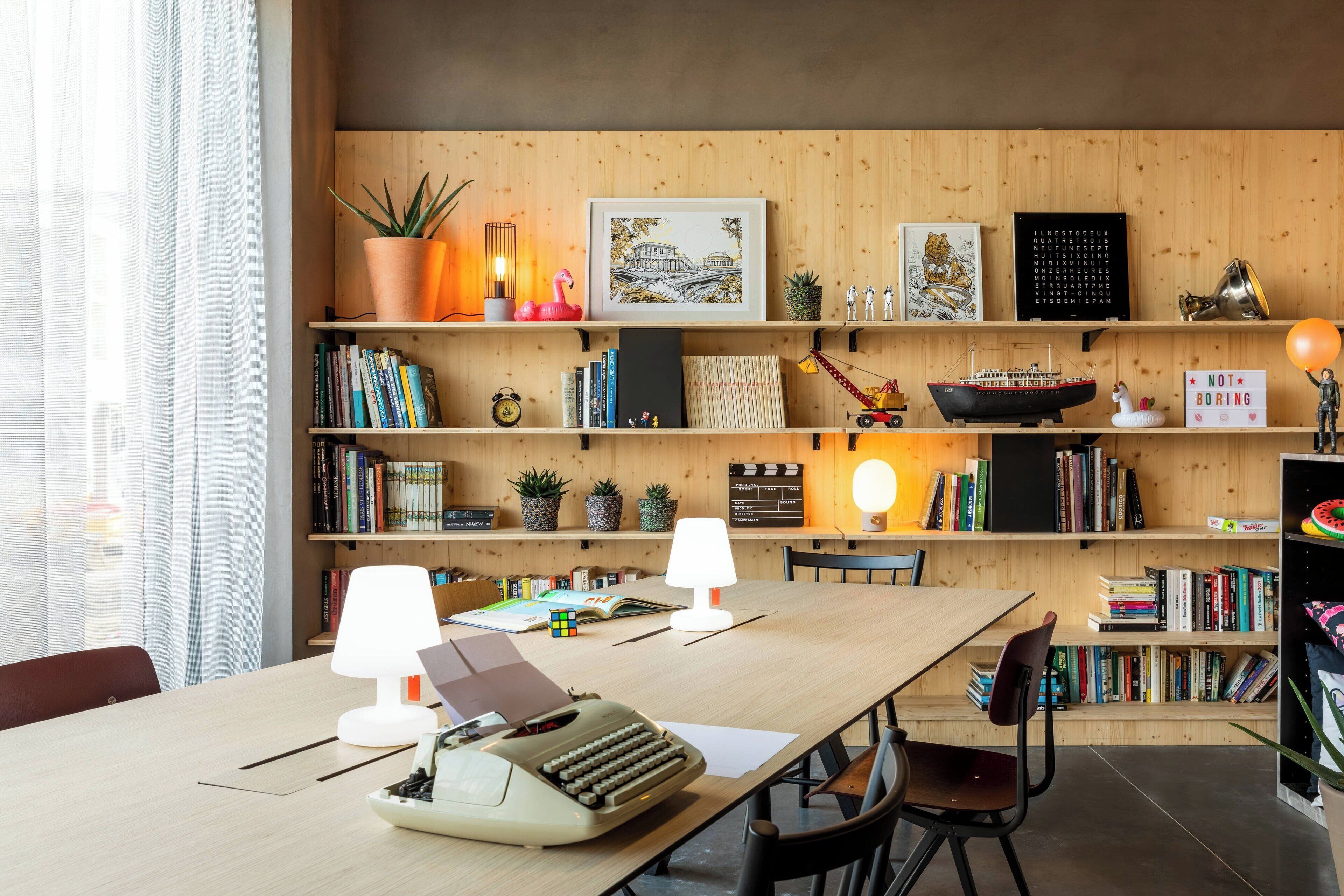 A modern, well-organized workspace with a wooden desk, two lamps, a vintage typewriter, and a bookshelf filled with books and decorative items.