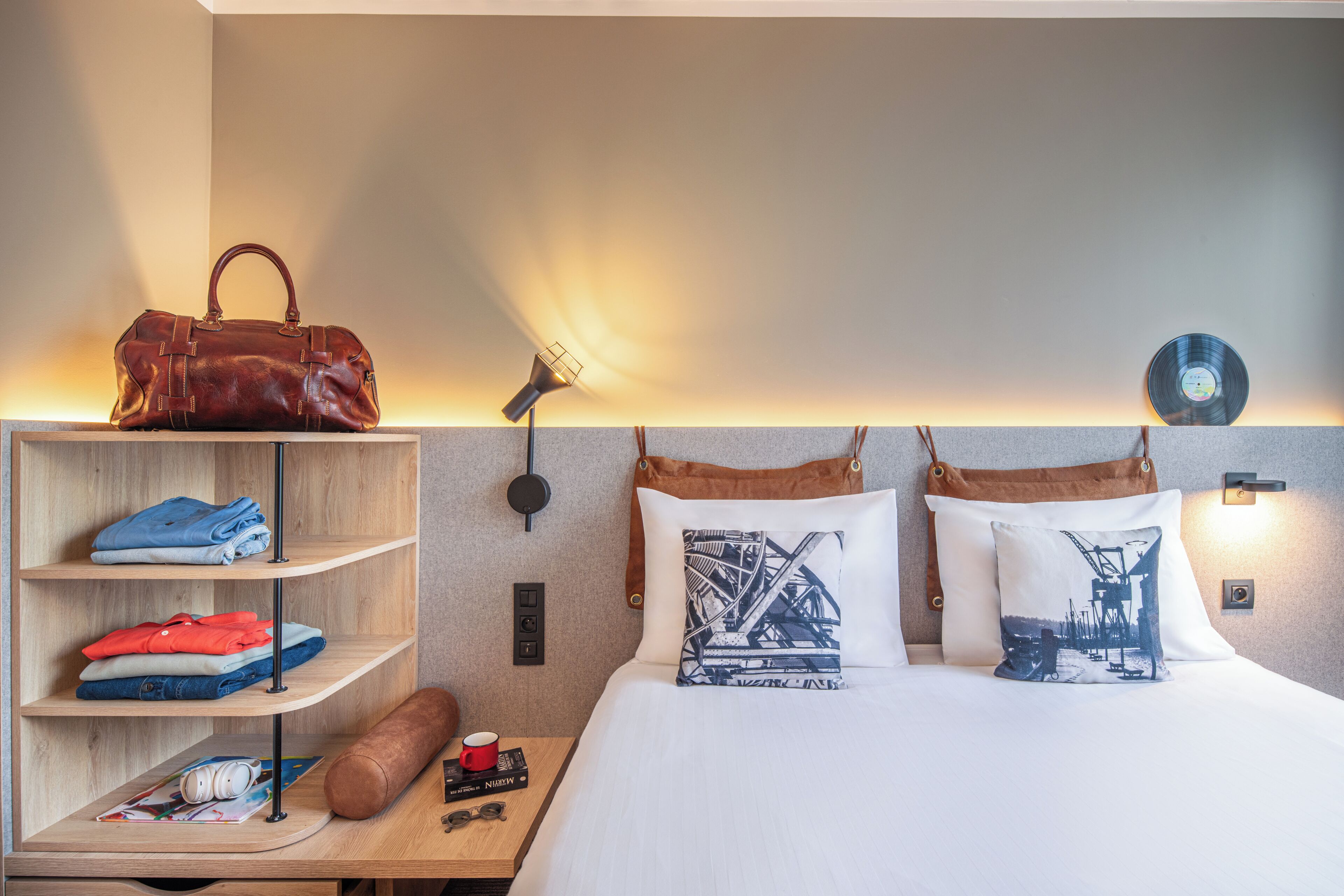 Una habitación de hotel con una cama, un estante con ropa doblada, una taza roja, un bolso marrón y una lámpara fijada a la pared.