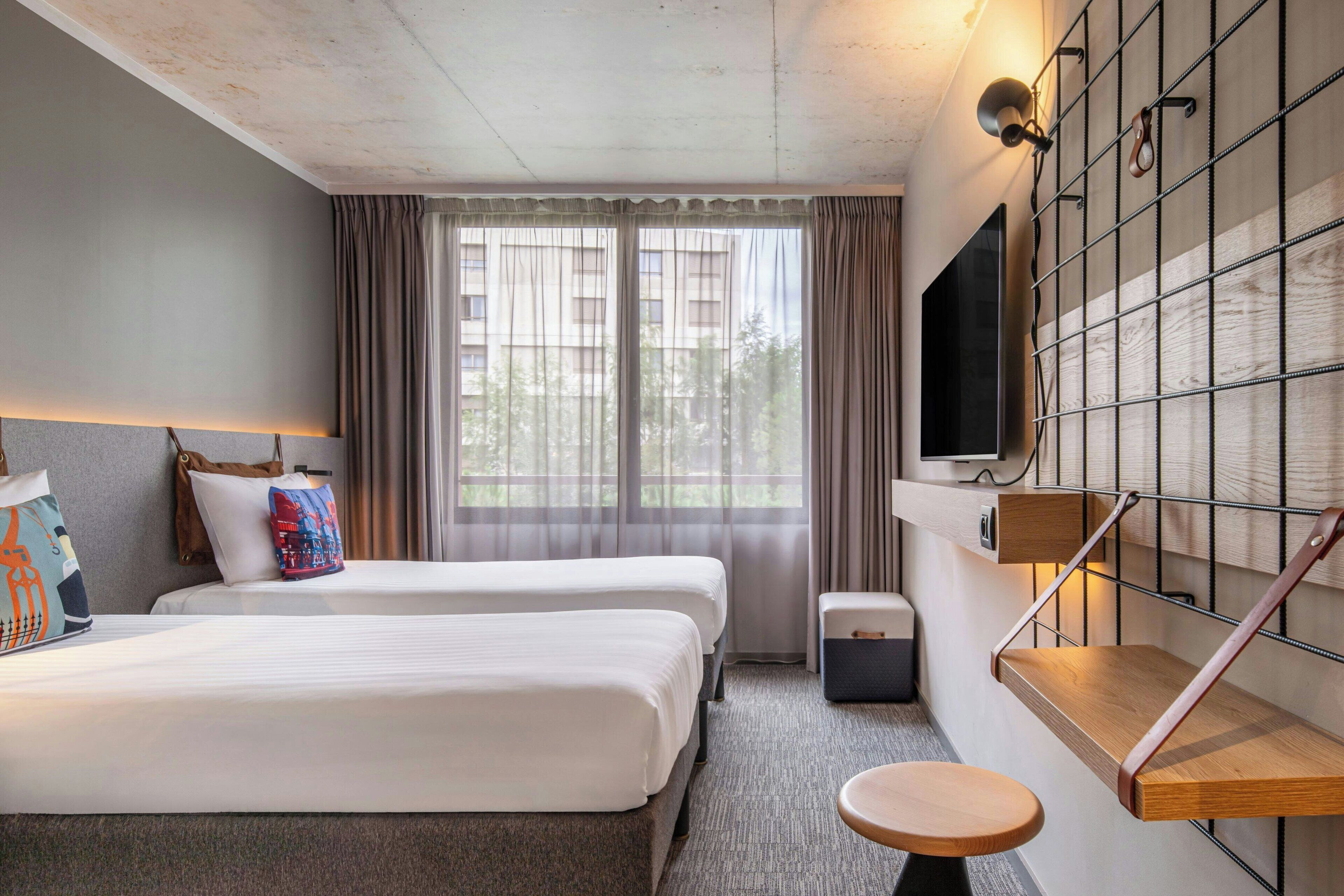 A hotel room with two beds, a small table, a TV mounted on the wall, and a window with curtains.