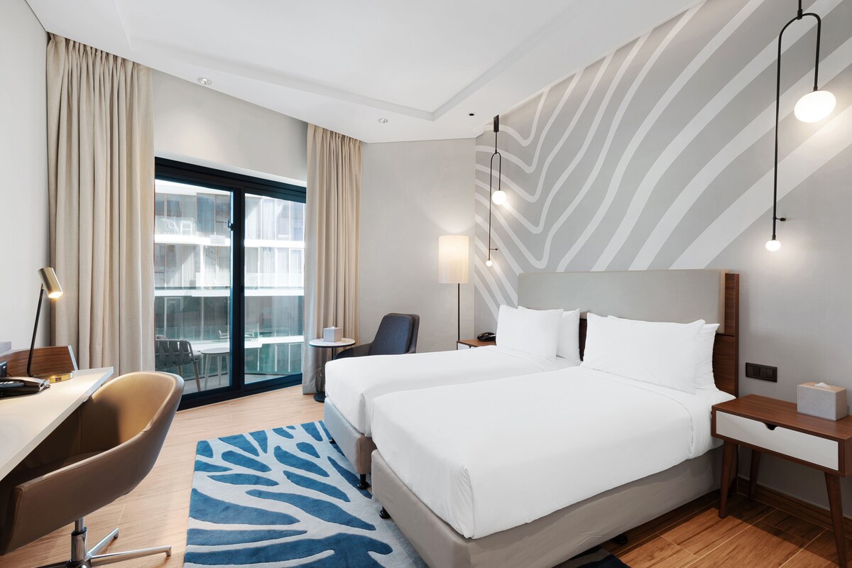 Habitación de hotel con dos camas, un escritorio, una silla, una lámpara y vistas al exterior.