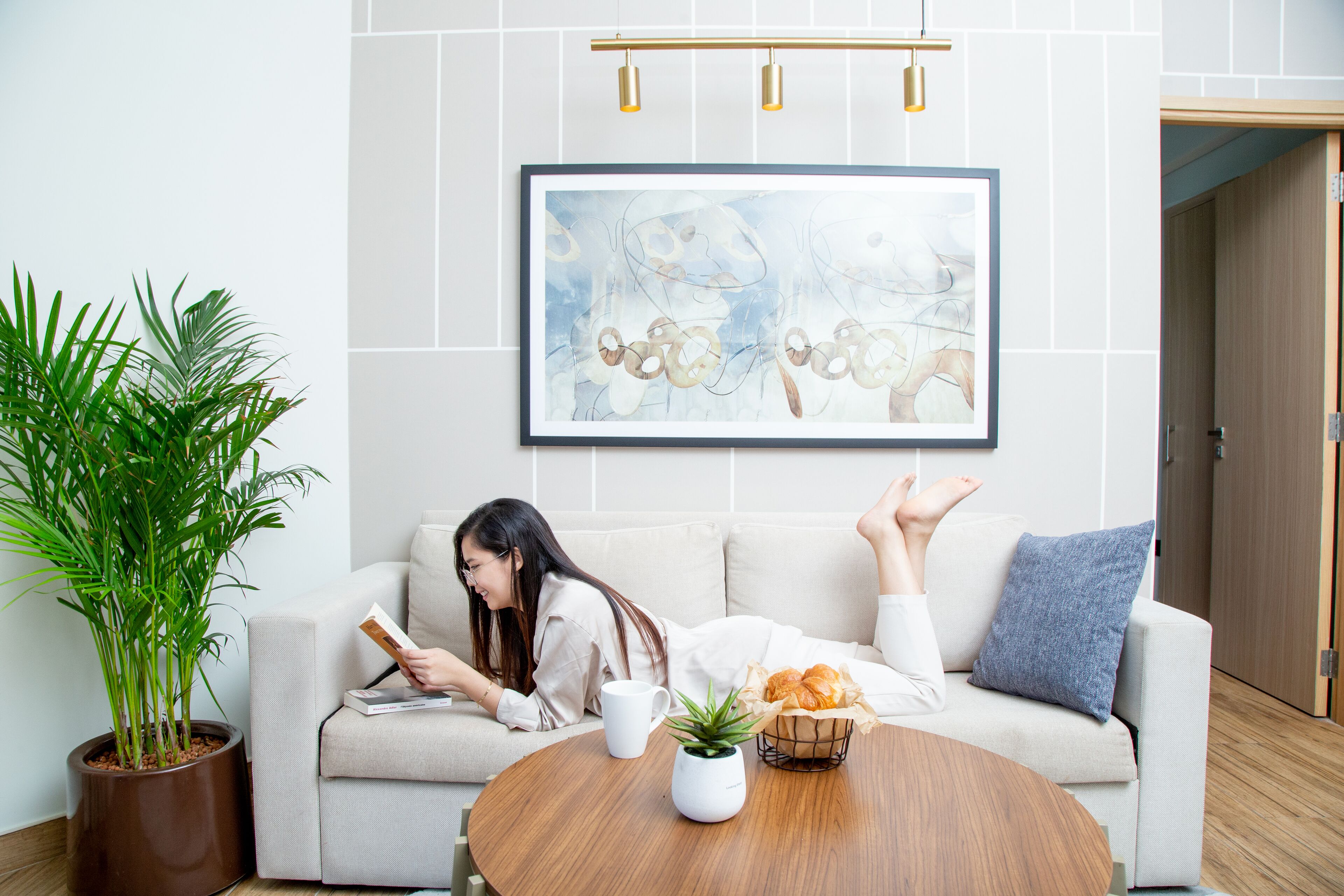 A woman reading on a sofa with a coffee table, a potted plant, and a framed artwork on the wall.