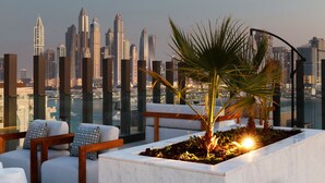 Un lounge en la azotea con vistas al horizonte urbano, muebles modernos y una jardinera con palmeras.