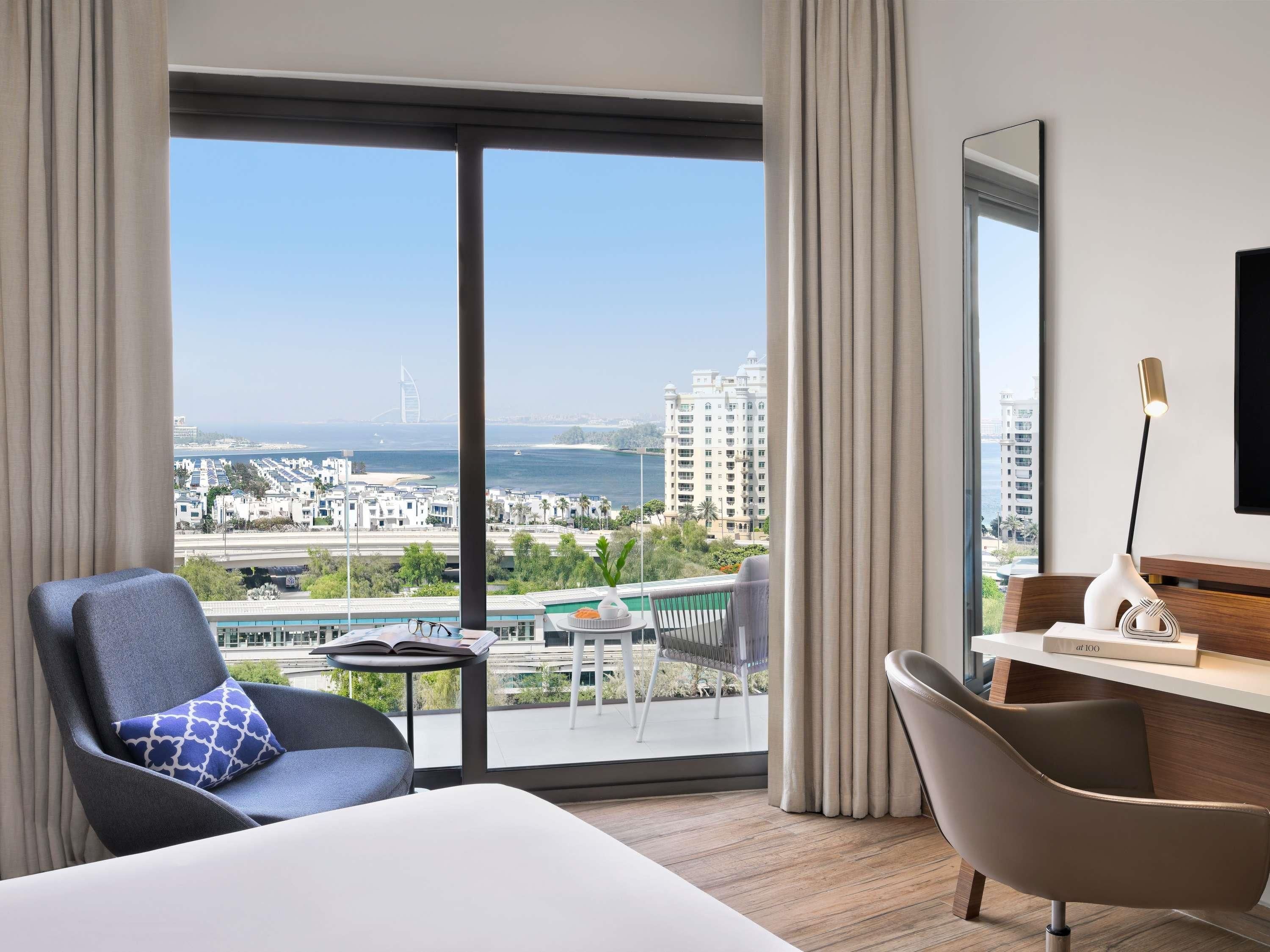 A hotel room with a large window, a bed, a desk, a chair, and a view of the city and sea.