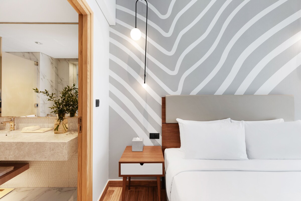 Una habitación de hotel moderna con baño de mármol, una mesita de noche de madera y una cama con ropa de cama blanca.
