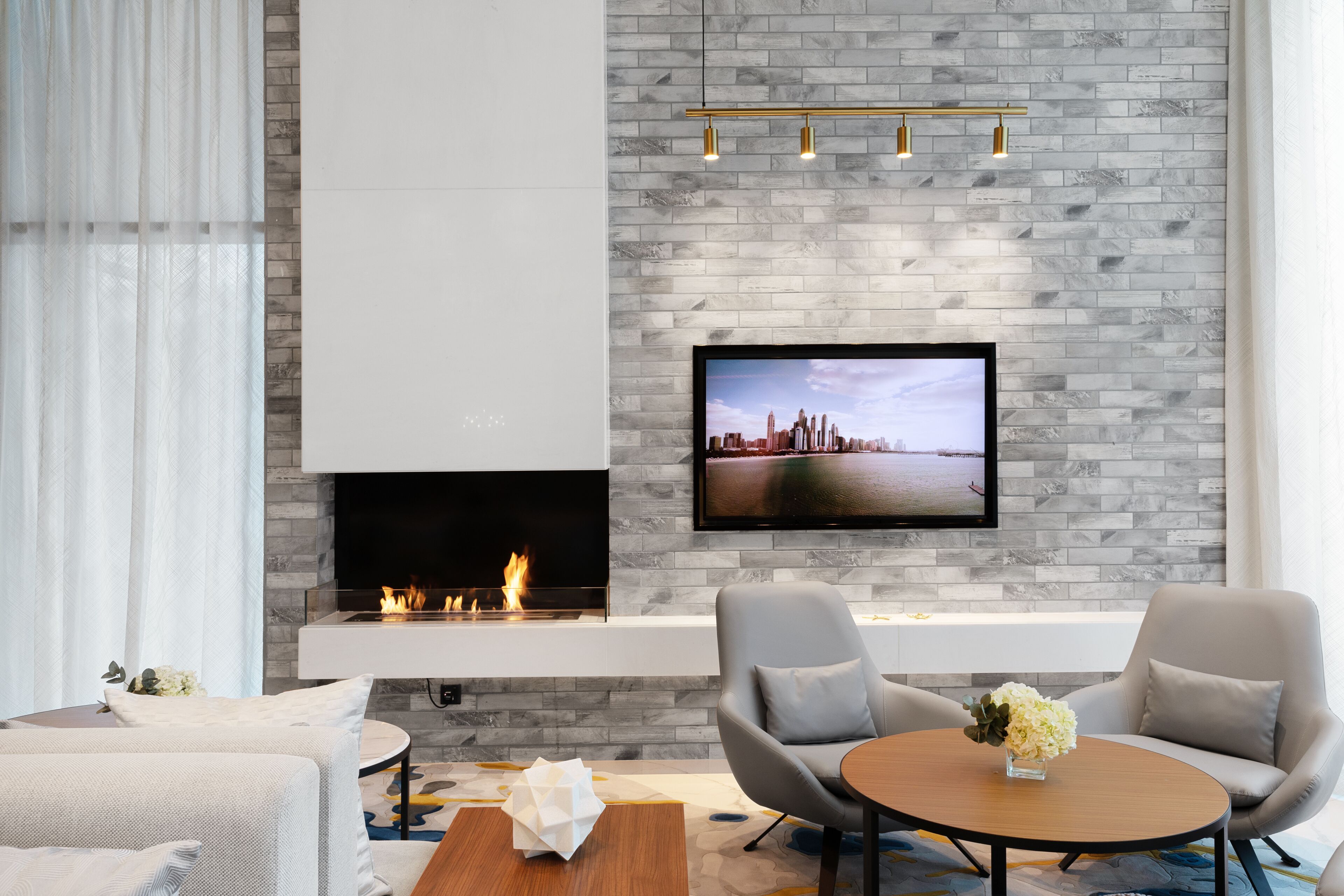 A modern living room with a fireplace, a flat-screen TV mounted on a brick wall, and two grey armchairs.