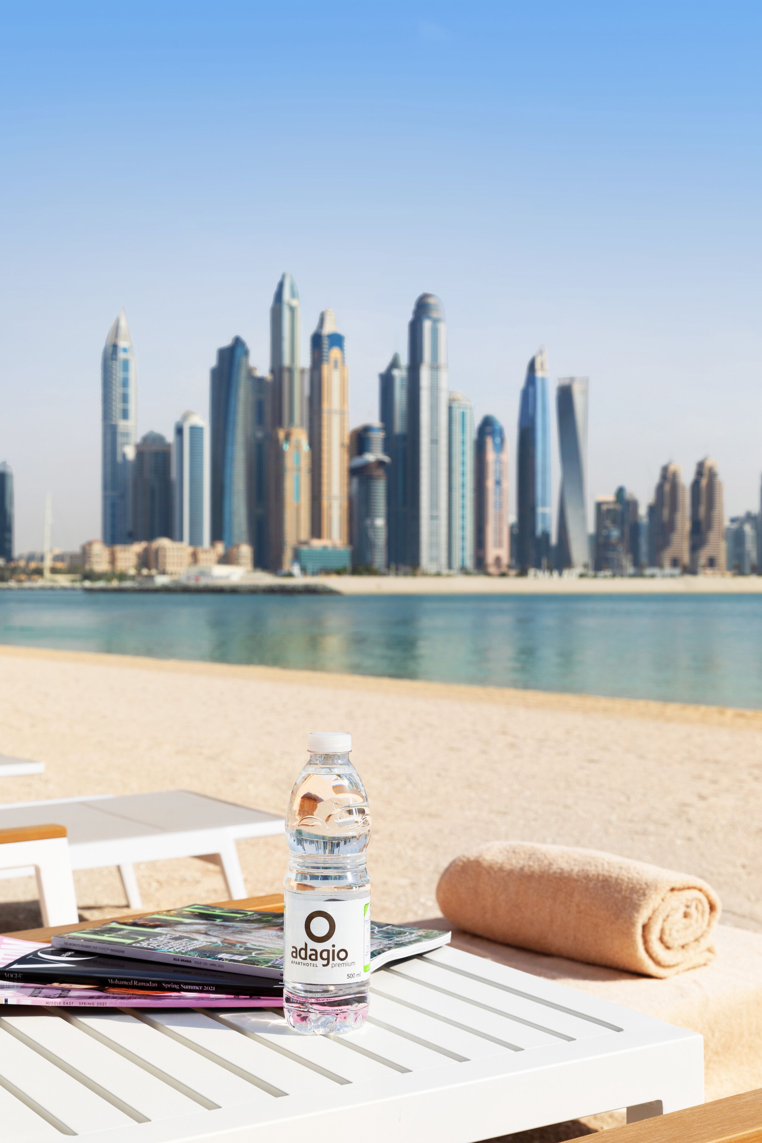 Una mesa con una botella de agua, una toalla enrollada y una revista en una playa, con un horizonte urbano al fondo.