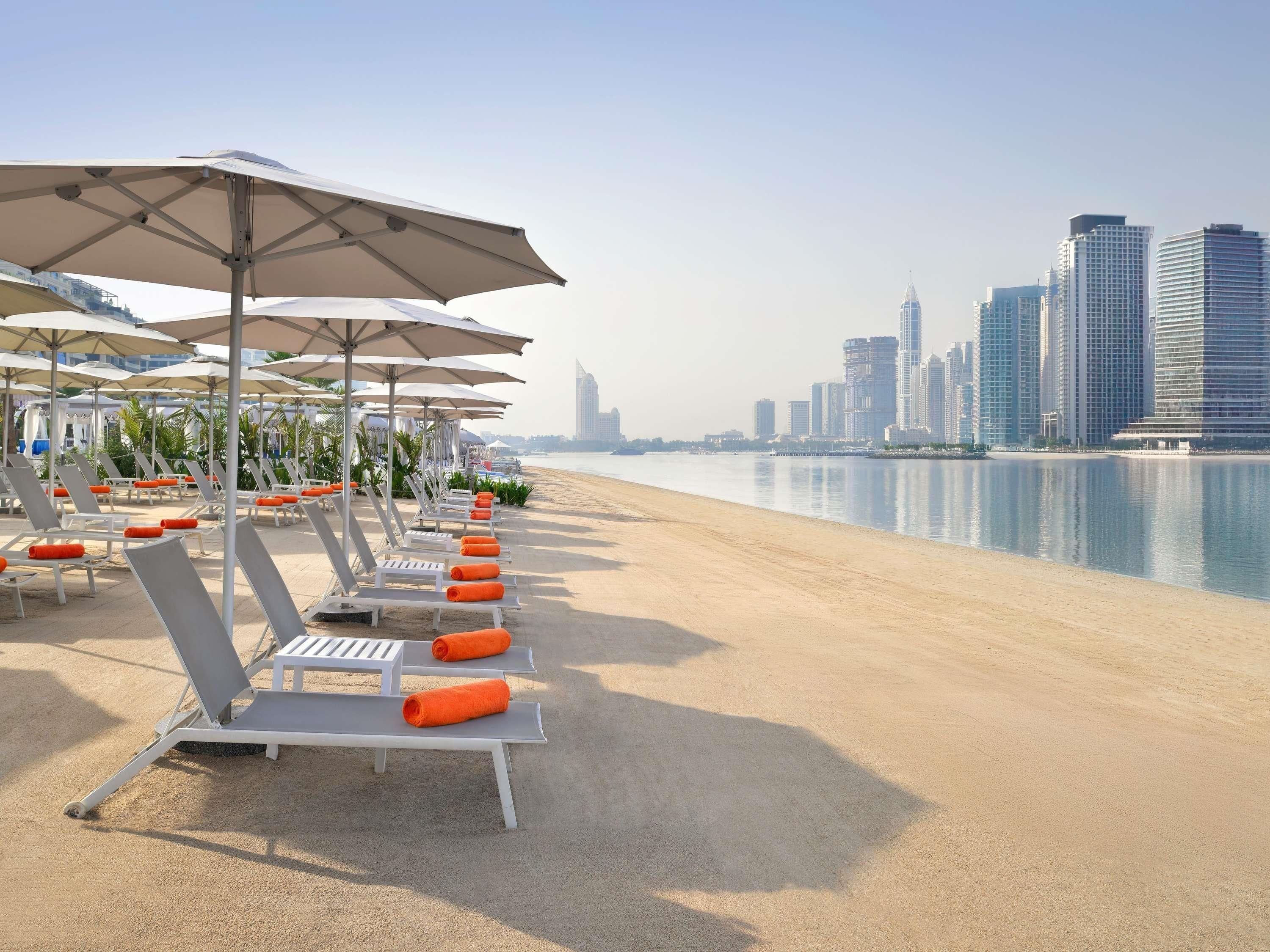Un hôtel situé en bord de mer, doté de chaises longues et de parasols, avec une plage de sable et, en arrière-plan, la silhouette de la ville.