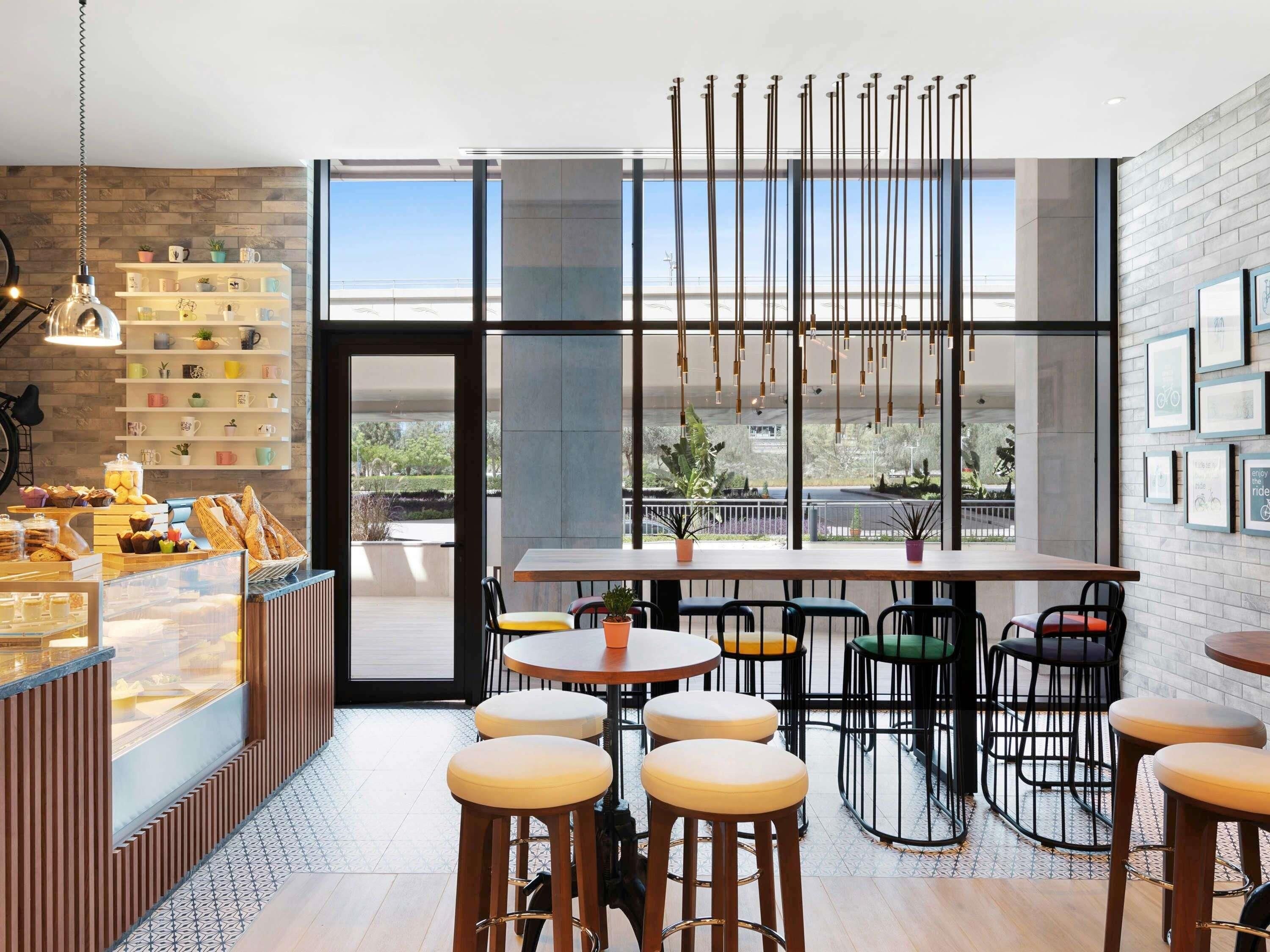 Una cafetería de estilo moderno, con una barra donde se ofrecen pasteles y bebidas, diversas opciones de asientos y grandes ventanales con vistas al exterior.
