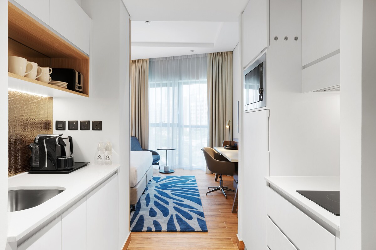 Una habitación de hotel moderna con kitchenette, una cama, un escritorio y un ventanal con cortinas.