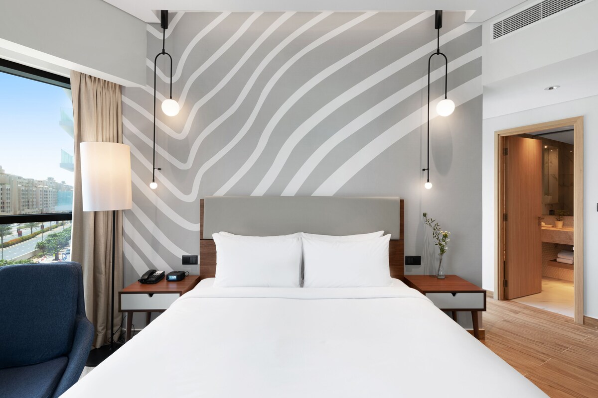 Habitación de hotel con una cama grande, un escritorio con lámpara, una silla y vistas a la ciudad a través de la ventana.