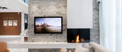 A modern living room with a fireplace, a flat-screen TV mounted on a brick wall, and a sofa with cushions.