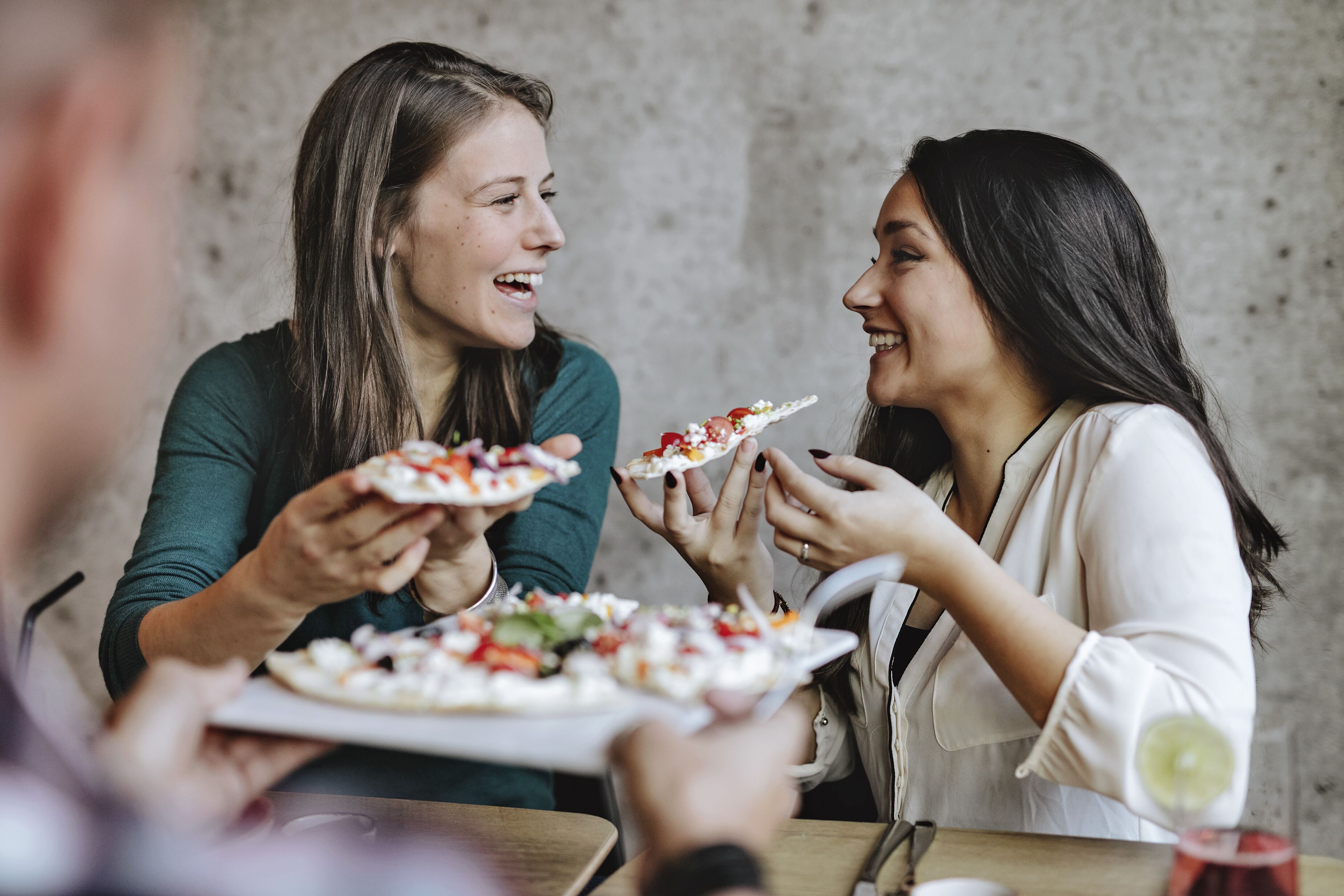 To personer holder tallerkener med mad, sandsynligvis pizza, i en afslappet spisesituation med en betonvæg i baggrunden.