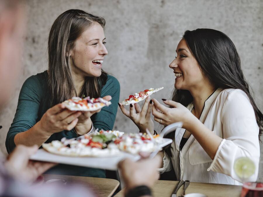 Dos personas sostienen platos con comida, probablemente pizza, en un ambiente de comedor informal con una pared de concreto al fondo.