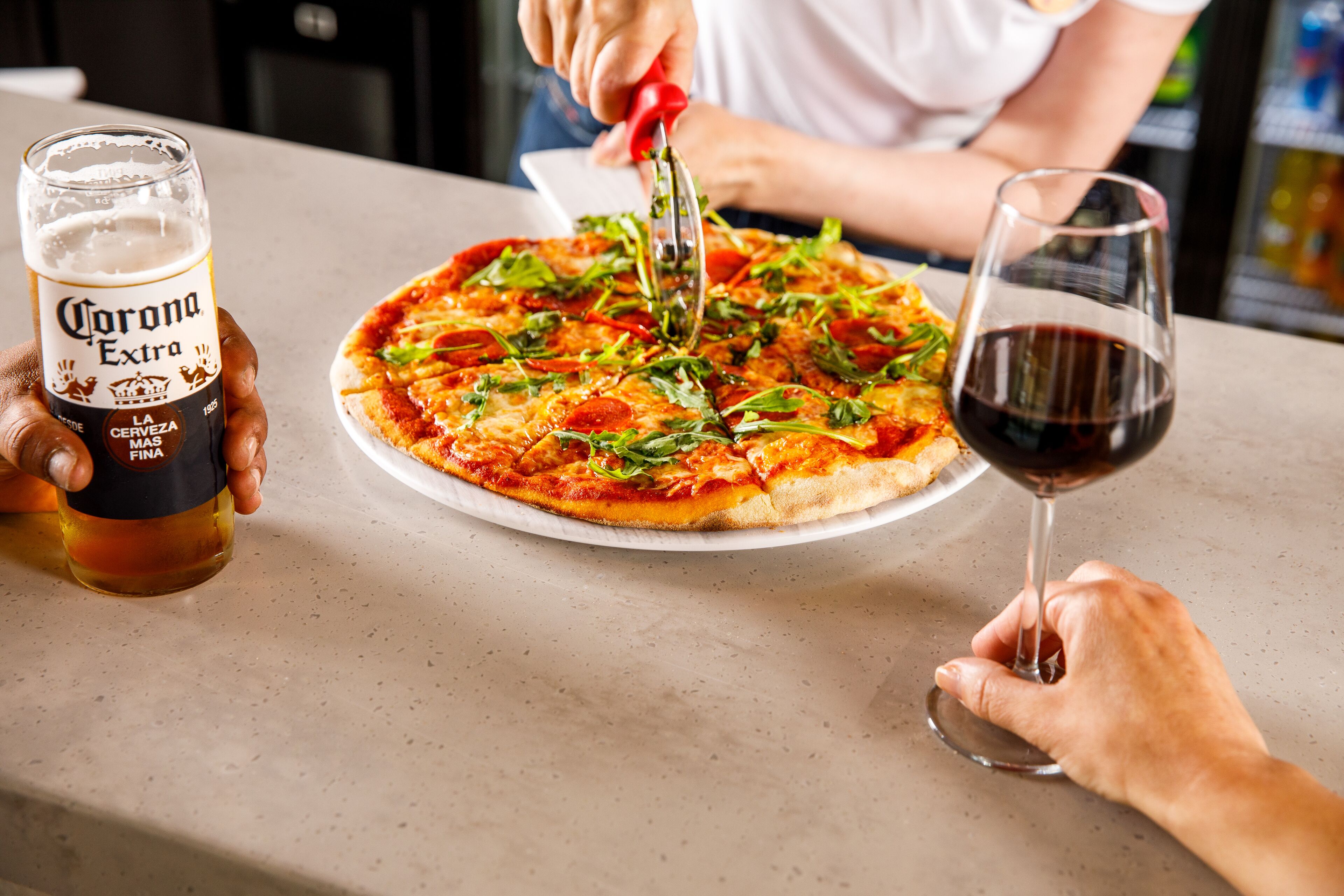 A person cutting a pizza with a pizza cutter on a table. There is a glass of Corona Extra beer and a glass of red wine on the table.