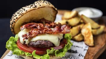 Hamburger con bacon, formaggio, lattuga, pomodoro e pane ai semi di sesamo, servito con patatine fritte su un tagliere di legno.