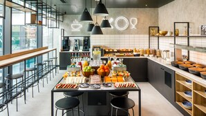 A modern cafe with a long communal table, bar seating, and a variety of food and drink options. - ibis London Sutton Point (Sutton)