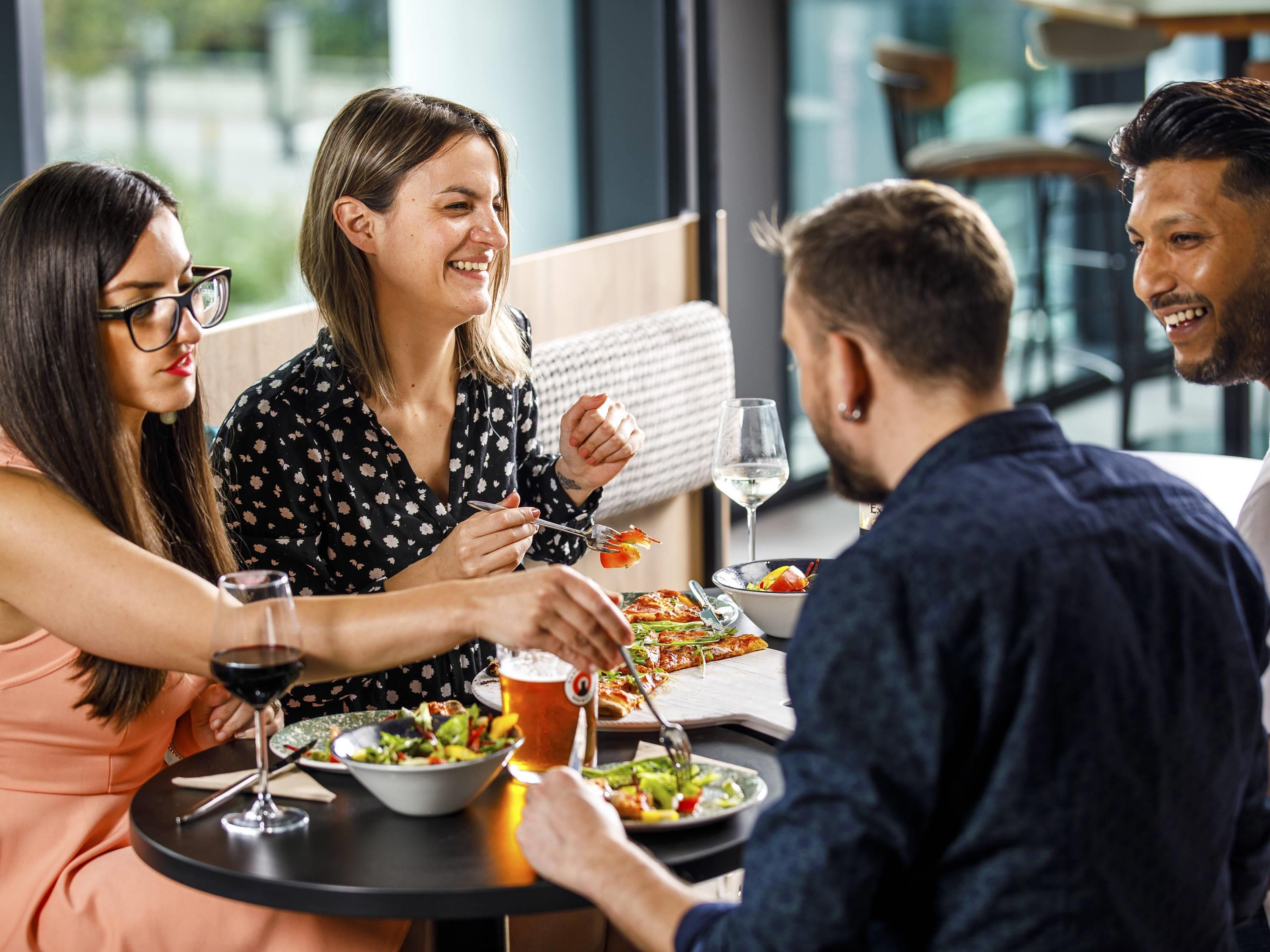 Un grupo de personas cenando en una mesa con varios platos, bebidas y una copa de vino.