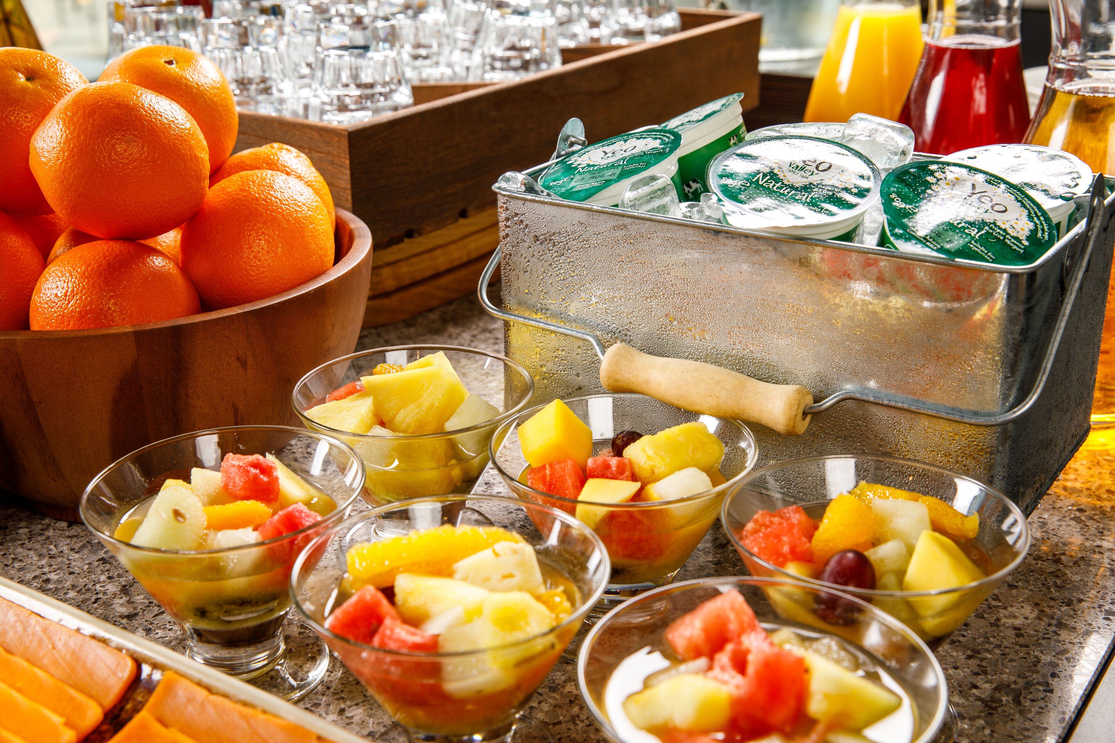 A breakfast buffet with bowls of fruit, a bowl of oranges, and a cooler with beverages.