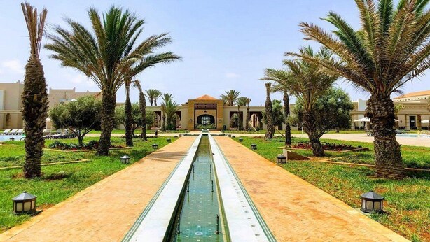 Un jardín simétrico con una fuente central, palmeras y un edificio con arcos al fondo.
