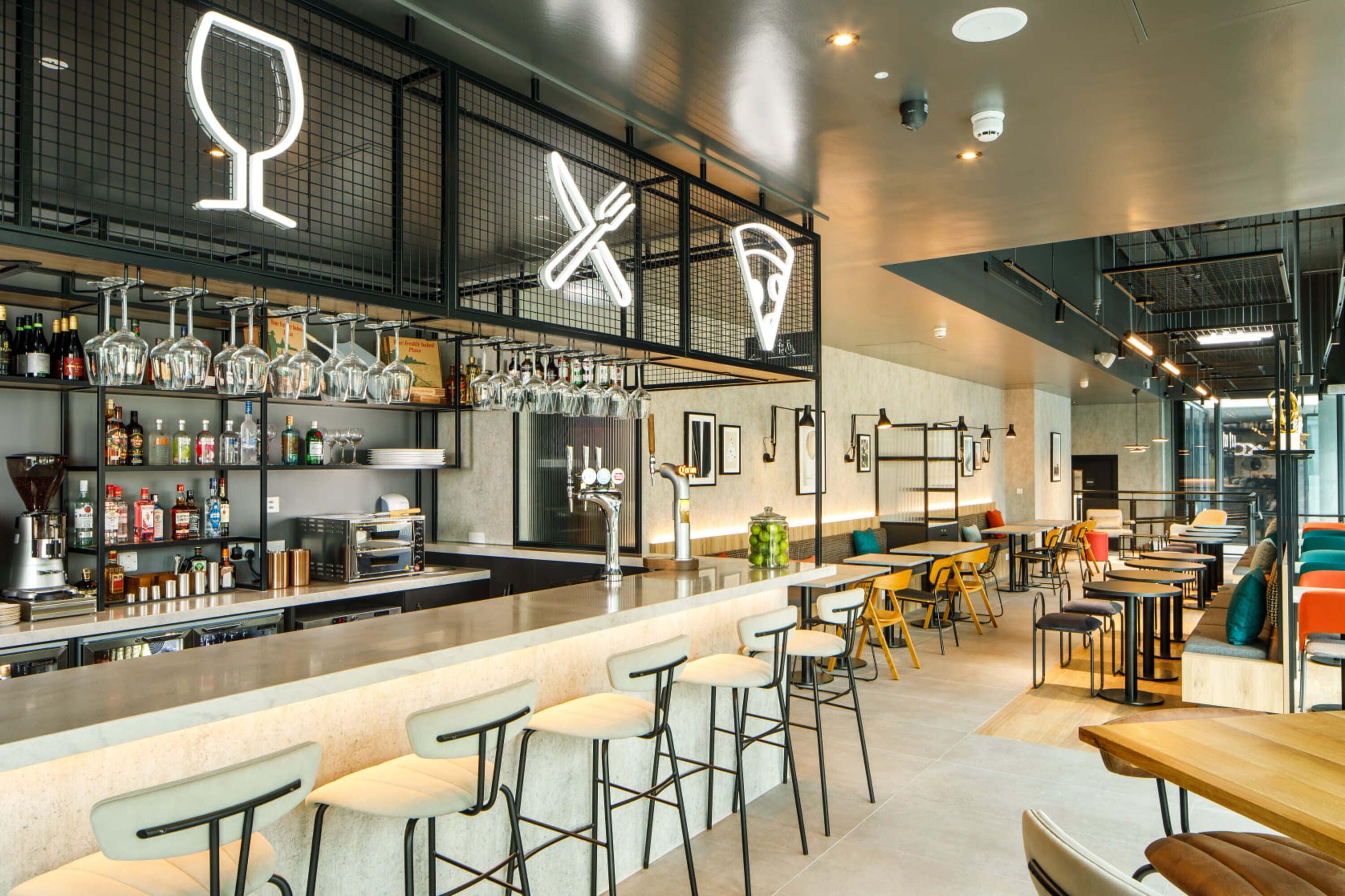 Zona de bar moderna con taburetes, una barra y estanterías llenas de botellas y vasos.