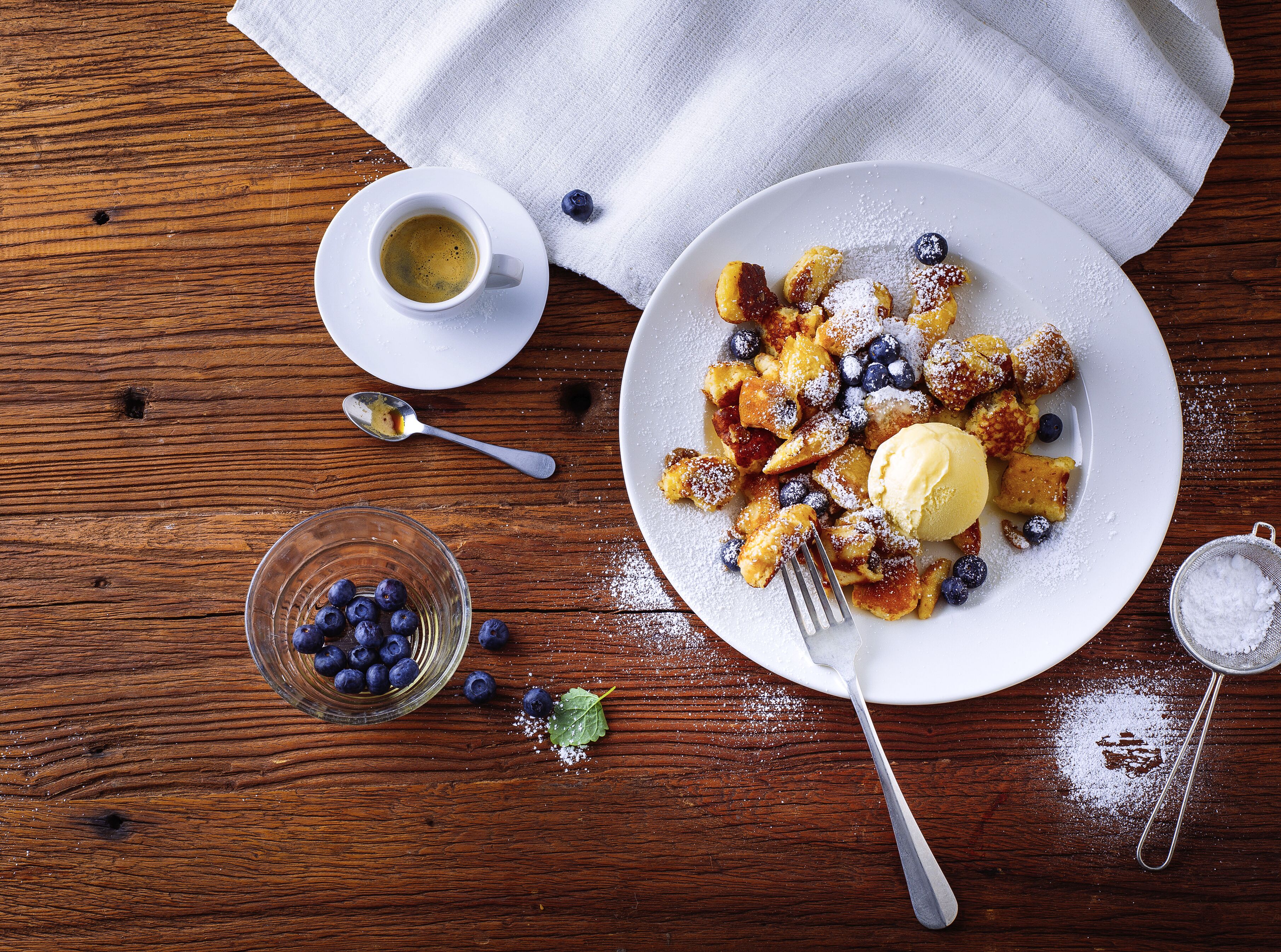 A wooden table with a plate of food, a glass of blueberries, and a cup of coffee.