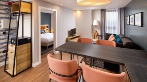 Ein modernes Hotelzimmer mit Holzboden, einer Essecke mit dunklem Tisch und Pfirsichfarbenen Stühlen, einem Bett mit Kopfschild in Schwarzweißmuster und einem großen Fenster mit Vorhängen.