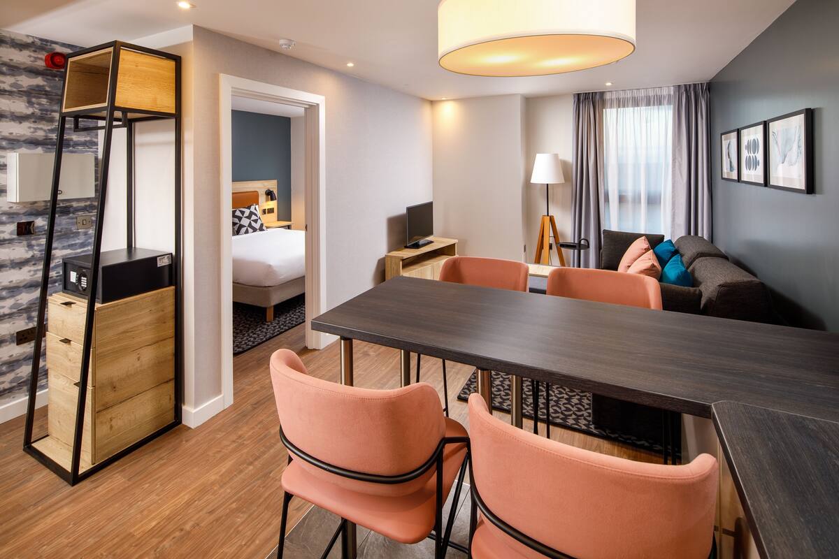 A modern hotel room with a wooden floor, a dining area with a dark table and peach chairs, a bed with a black and white patterned headboard, and a large window with curtains.