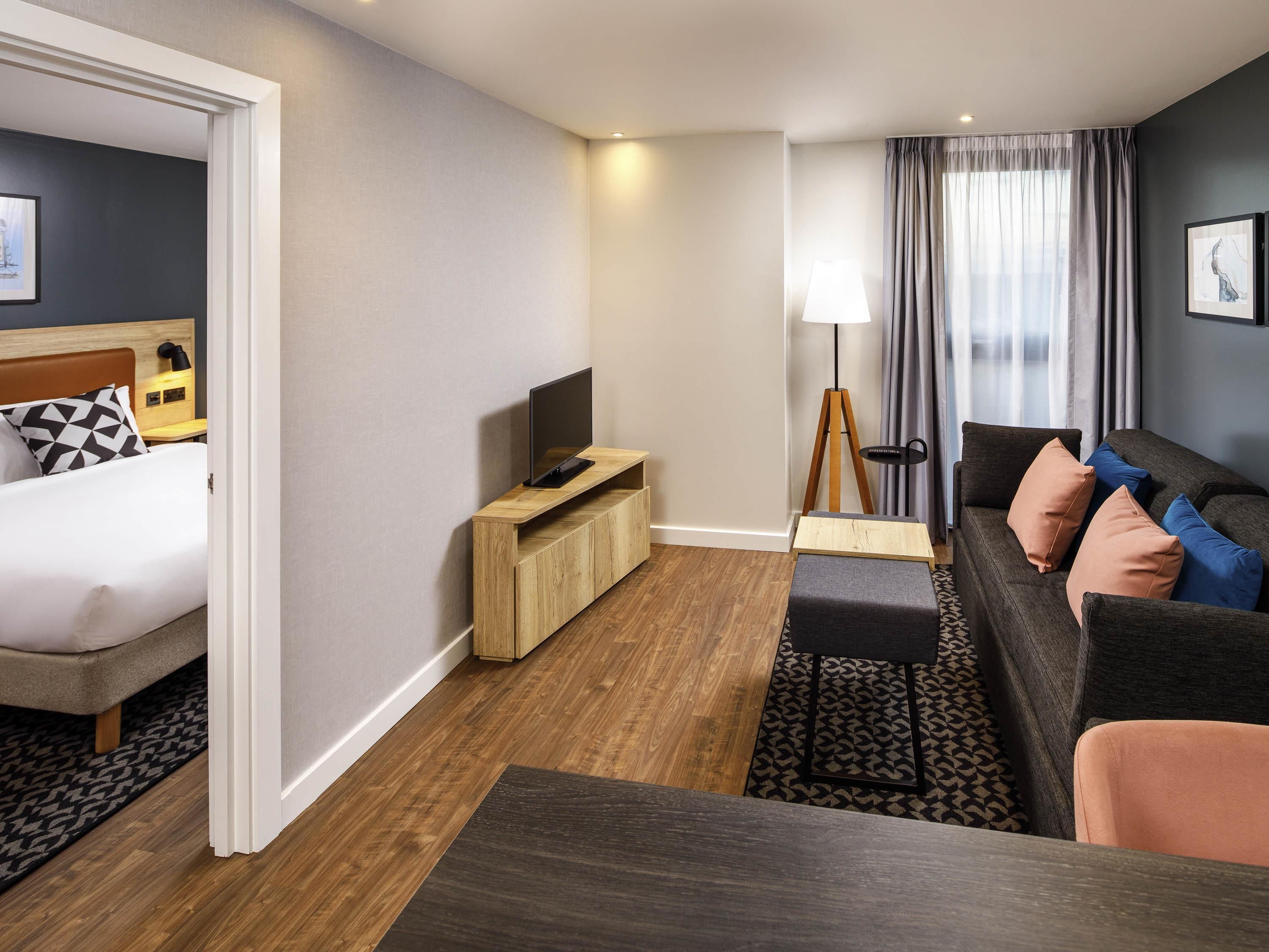 A hotel room with a bed, a TV on a wooden stand, a sofa with cushions, a floor lamp, and a window with curtains.