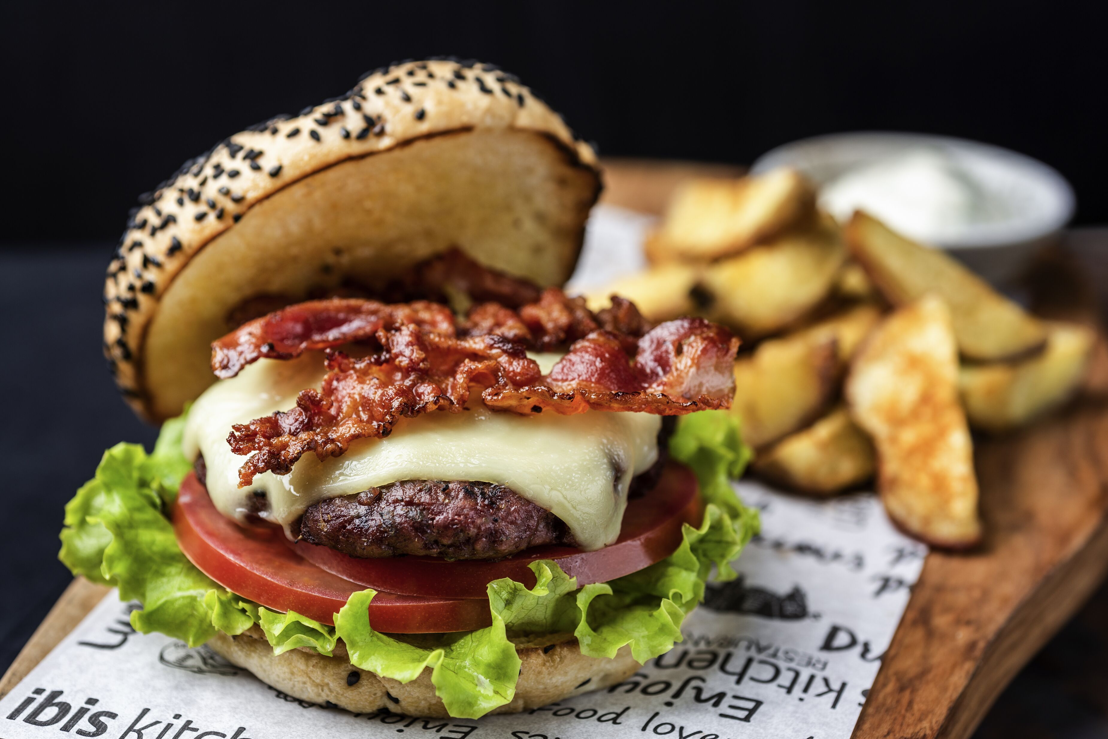 A burger with bacon, cheese, lettuce, tomato, and a sesame seed bun, served with fries on a wooden board.
