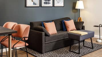 A modern living room with a grey sofa, orange chairs, a coffee table, and a patterned rug. There are three framed artworks on the wall.