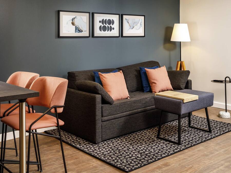 A modern living room with a grey sofa, orange chairs, a coffee table, and a patterned rug. There are three framed artworks on the wall.
