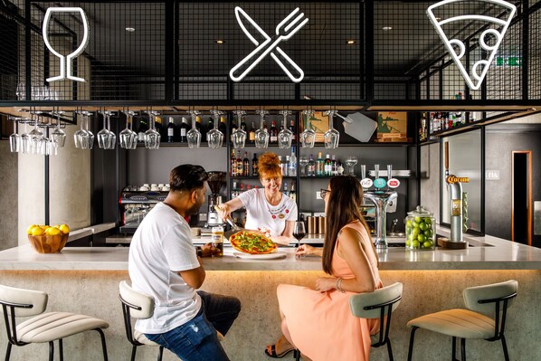 Zona de bar moderna con una barra, copas de vino colgantes y una pizza siendo servida.