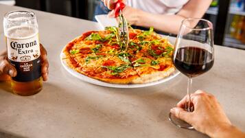 A person cutting a pizza with a pizza cutter on a table. There is a glass of Corona Extra beer and a glass of red wine on the table.