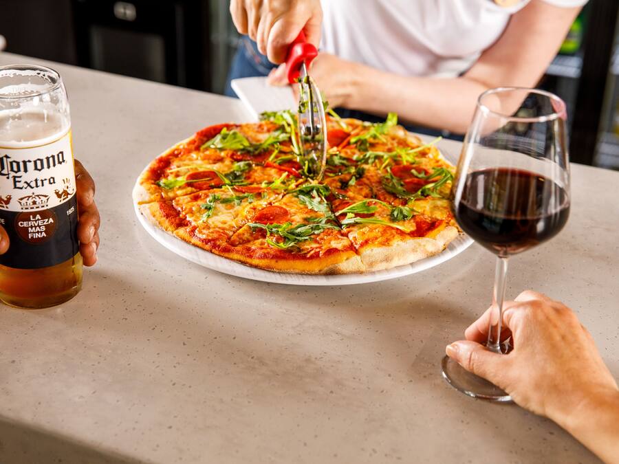 A person cutting a pizza with a pizza cutter on a table. There is a glass of Corona Extra beer and a glass of red wine on the table.