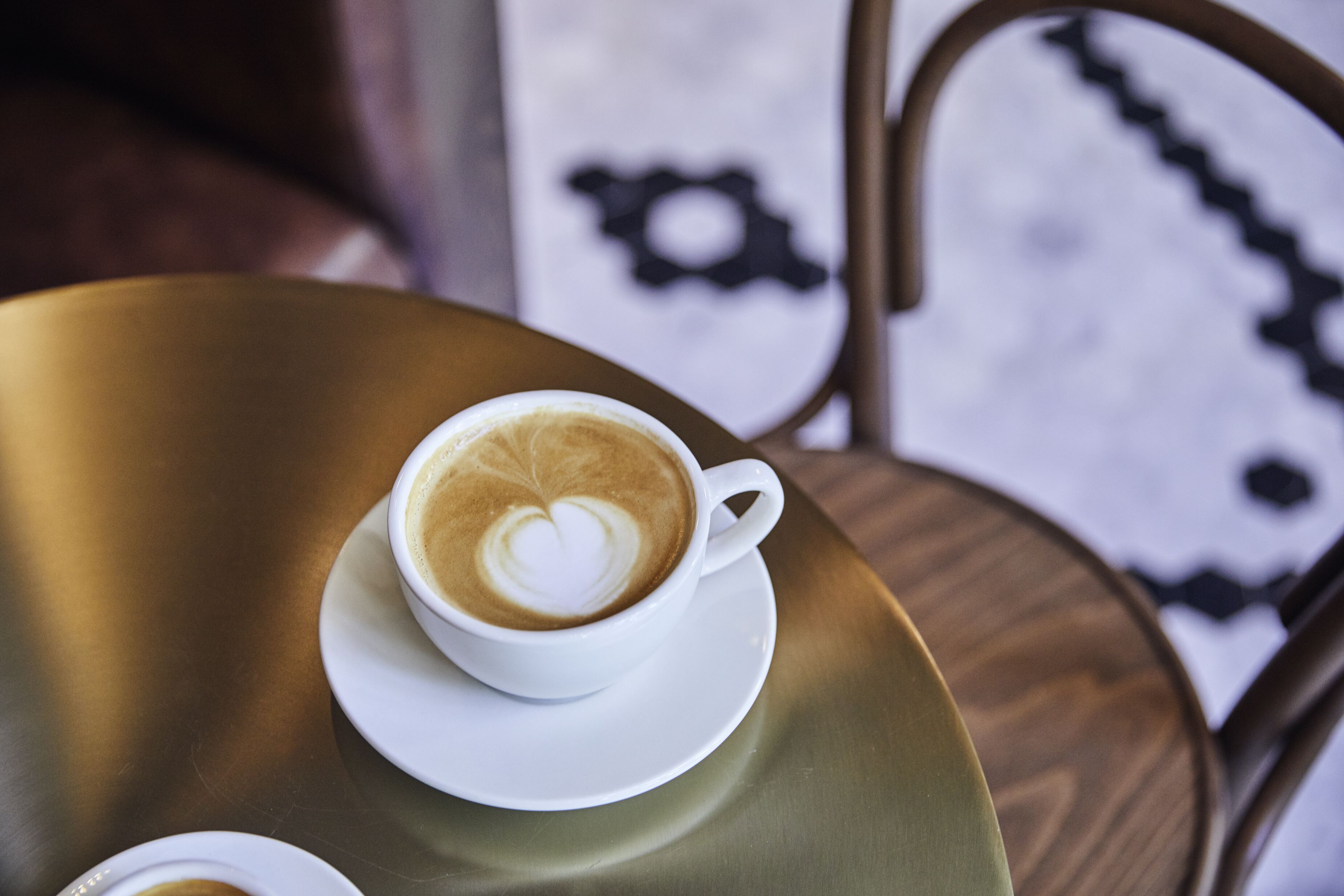 Una taza blanca de café con arte de latte sobre un platillo dorado, colocada sobre una mesa redonda.