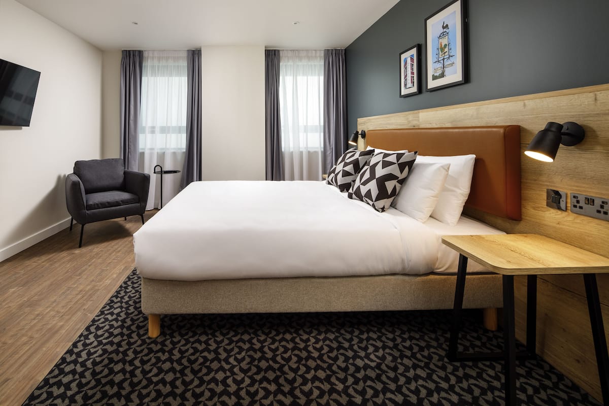 A modern hotel room with a large bed, a wooden nightstand, a chair, and a flat-screen TV mounted on the wall.