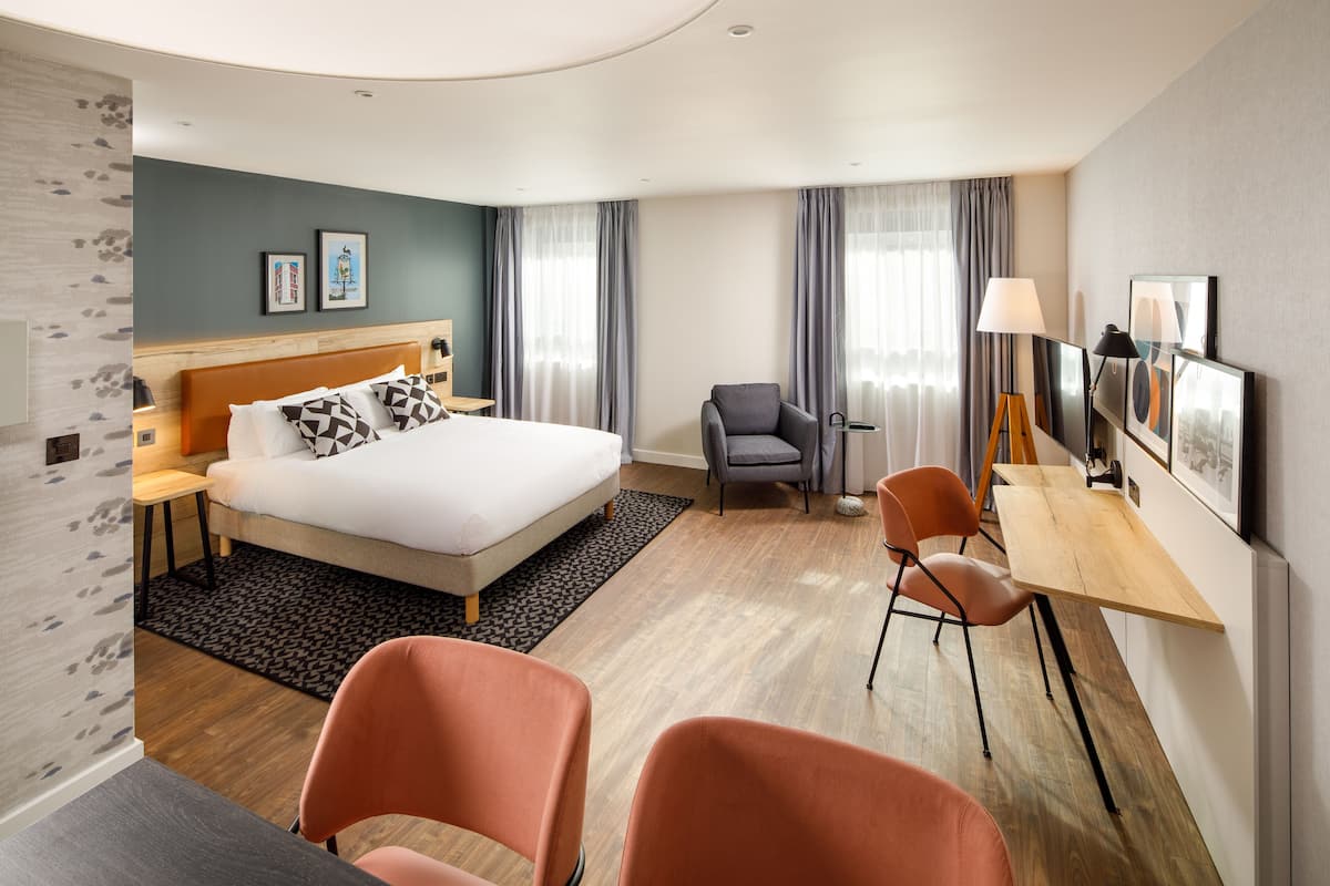 A hotel room with a bed, a desk, two chairs, a lamp, and artwork on the walls.