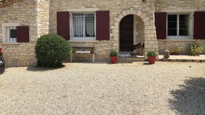 A stone building with a central entrance, two windows with shutters, a bench outside, and potted plants.