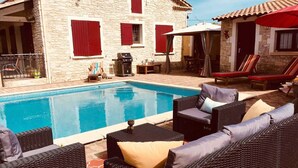 A stone building with red shutters, a swimming pool, wicker furniture, and a red umbrella.