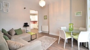 A modern living room with a glass dining table, white chairs, a beige sofa with green cushions, a chandelier, and a view into the kitchen.