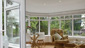 Een ruime kamer met een groot erkerraam, een eethoek met een glazen tafel en een comfortabele fauteuil.