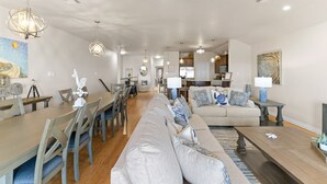 A spacious living area with a long sofa, dining table, and chairs. There is a kitchen in the background with cabinets and a microwave.