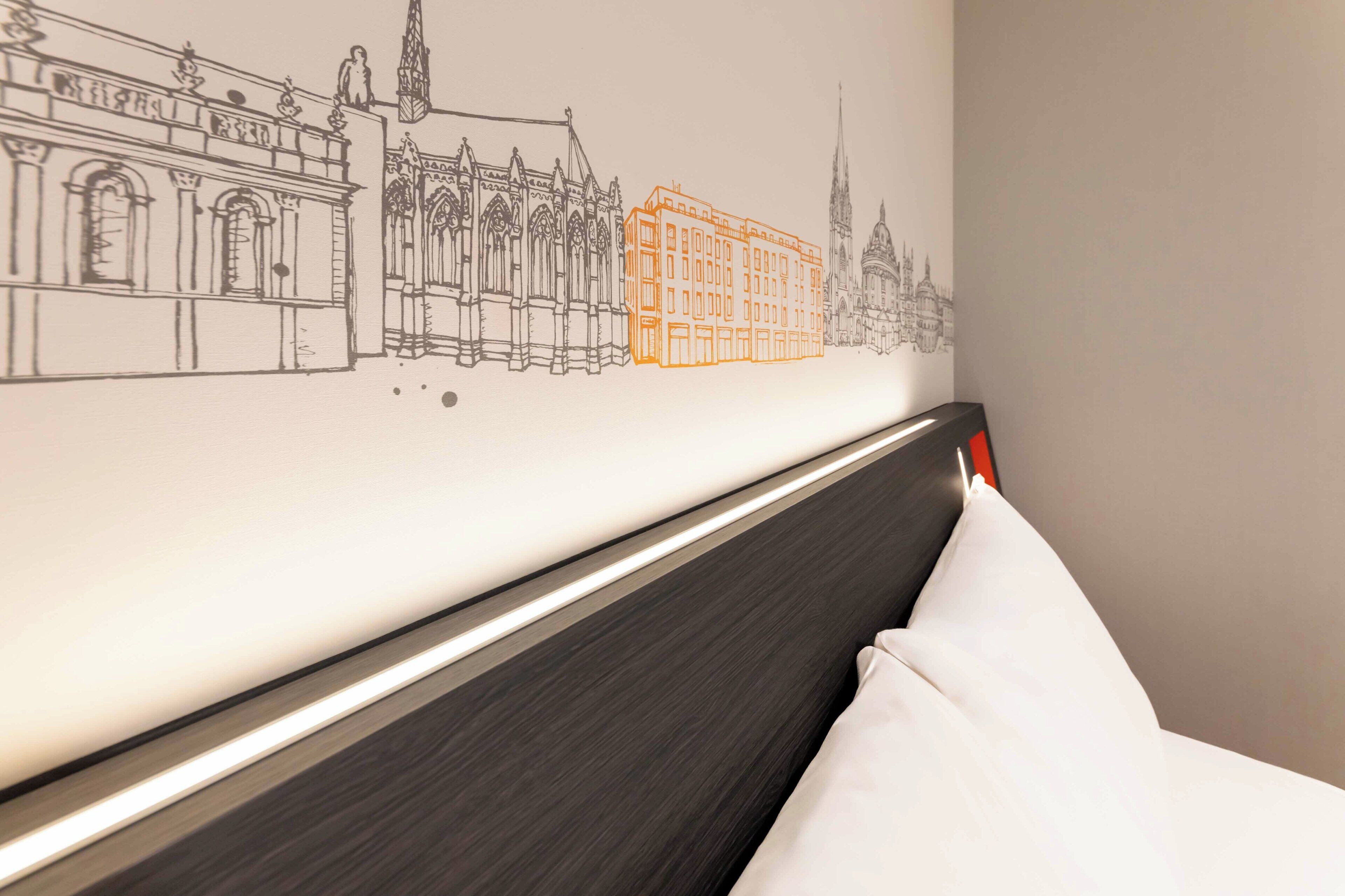 Quarto de hotel com cama, cabeceira e um mural na parede retratando uma paisagem urbana.