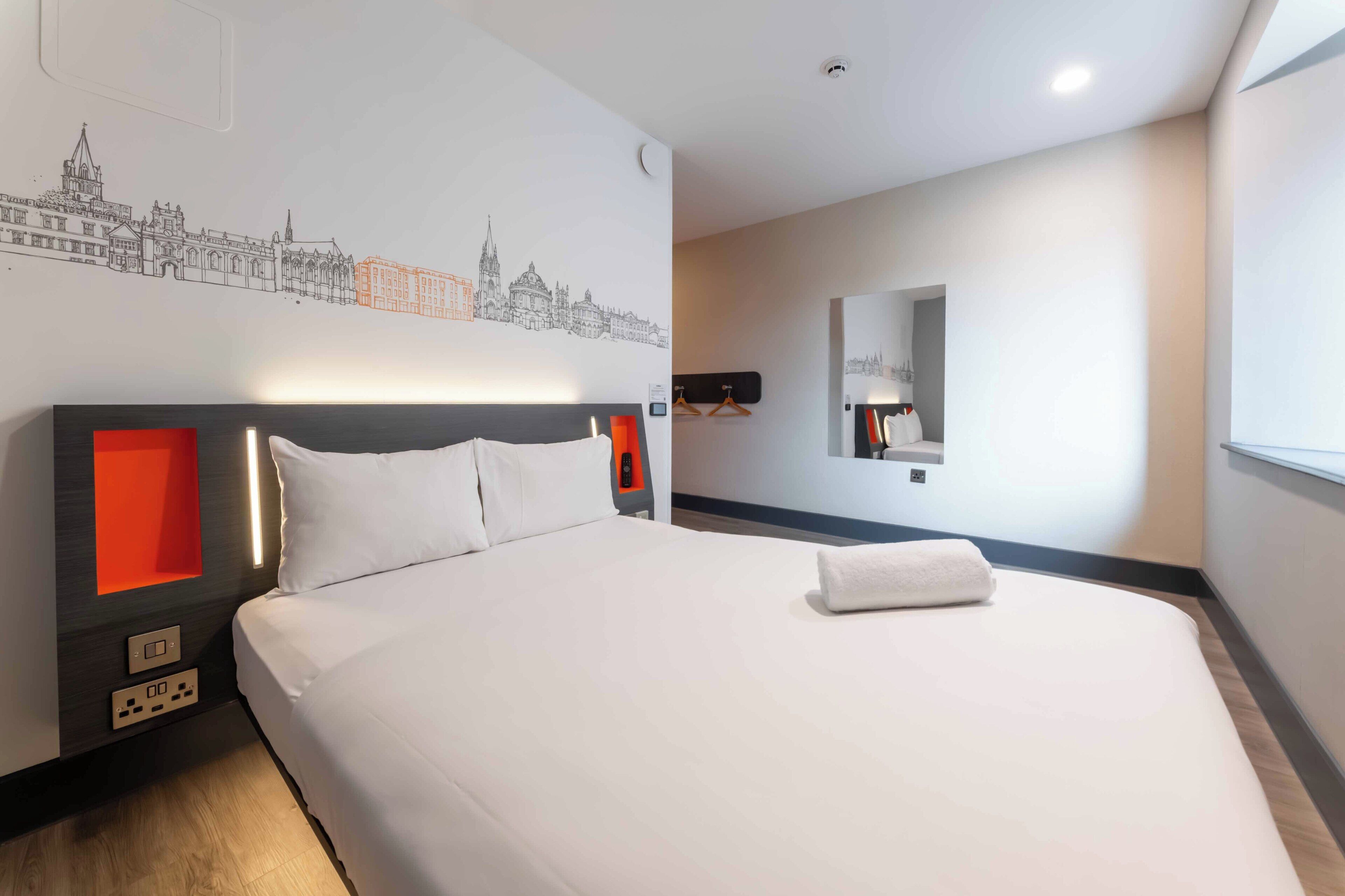 A hotel room with a bed, a wall-mounted mirror, and a cityscape artwork.