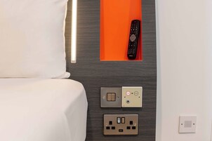 Habitación de hotel con cama, control remoto, interruptor de luz en la pared, termostato y enchufe.