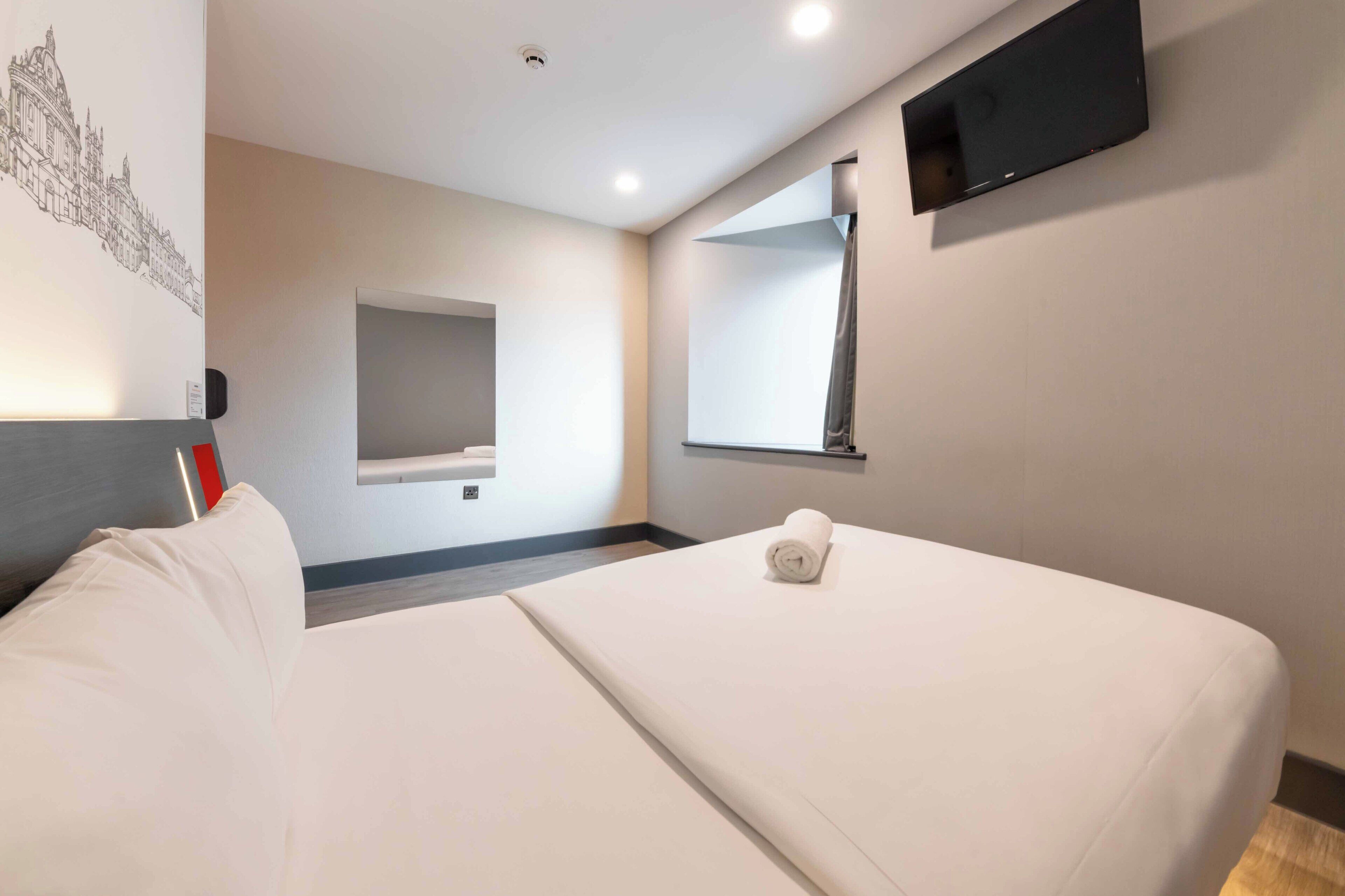A hotel room with a bed, a flat-screen TV, a wall-mounted mirror, and a wall art piece.